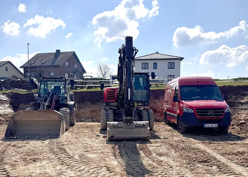 Vermietung Baumaschinen Bauplatz mit einem Radlader, einem Bagger und einem roten Mercedes-Transporter nebeneinander auf unbefestigtem Boden vor Wohnhäusern