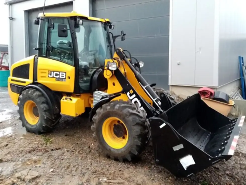 Gelber Radlader der Marke JCB mit schwarzer Schaufel auf matschigem Boden vor einer grauen Halle