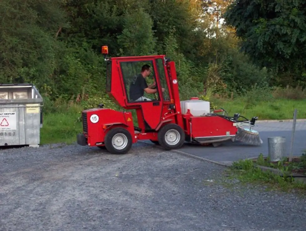 Roter Kehrmaschinen-Traktor mit Fahrer auf einem Schotterweg neben einem Metallmülleimer und einem Recycling-Container